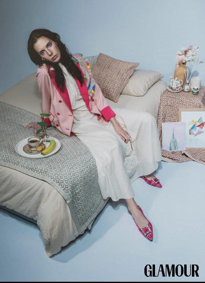 Woman sitting on a couch with a decorative setting, wearing a pink coat and white dress for Glamour Magazine.